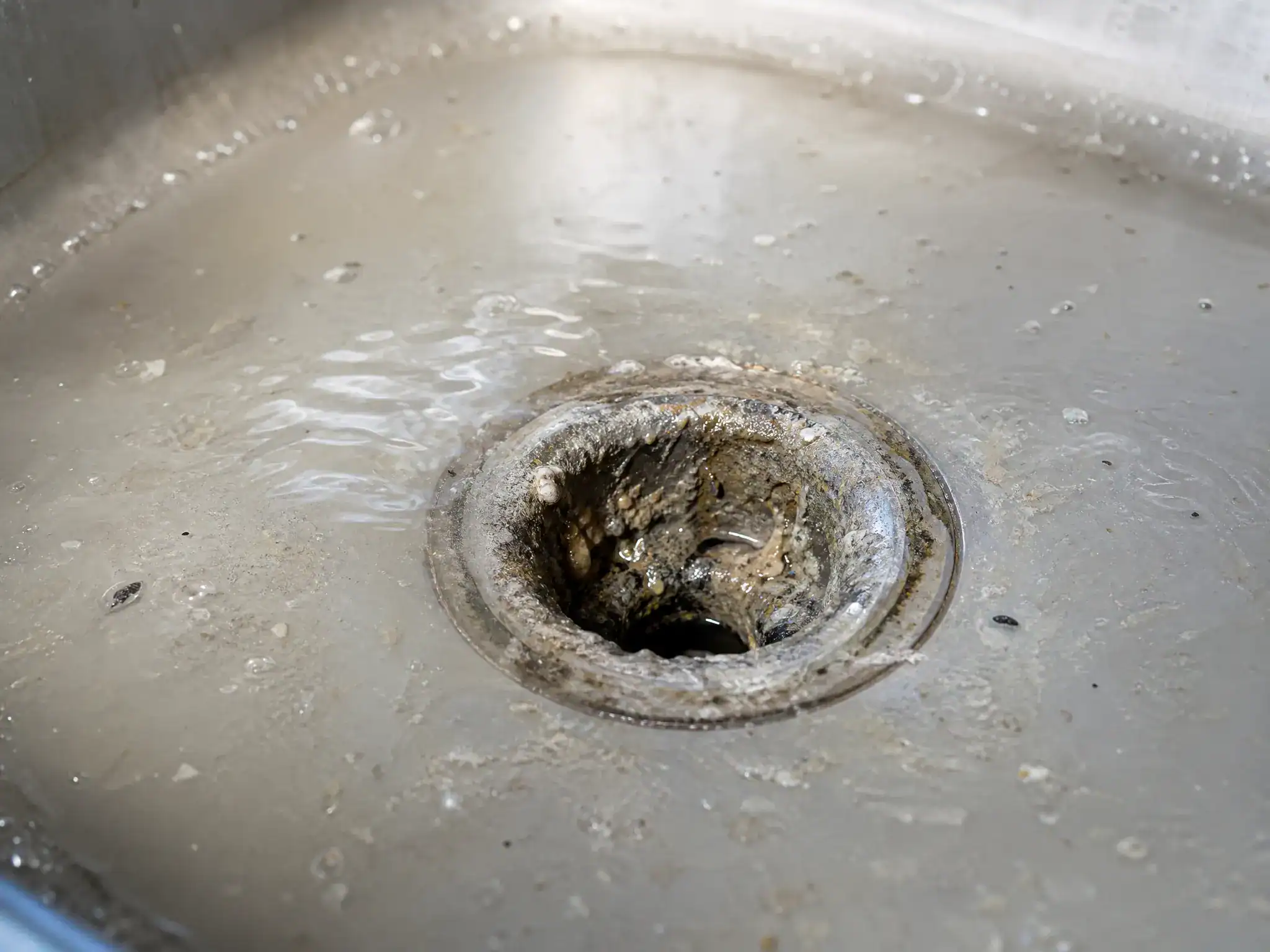 Close-up of a kitchen sink drain with cloudy, dirty water pooling around it and grime buildup visible on the drain, indicating a clogged sink.