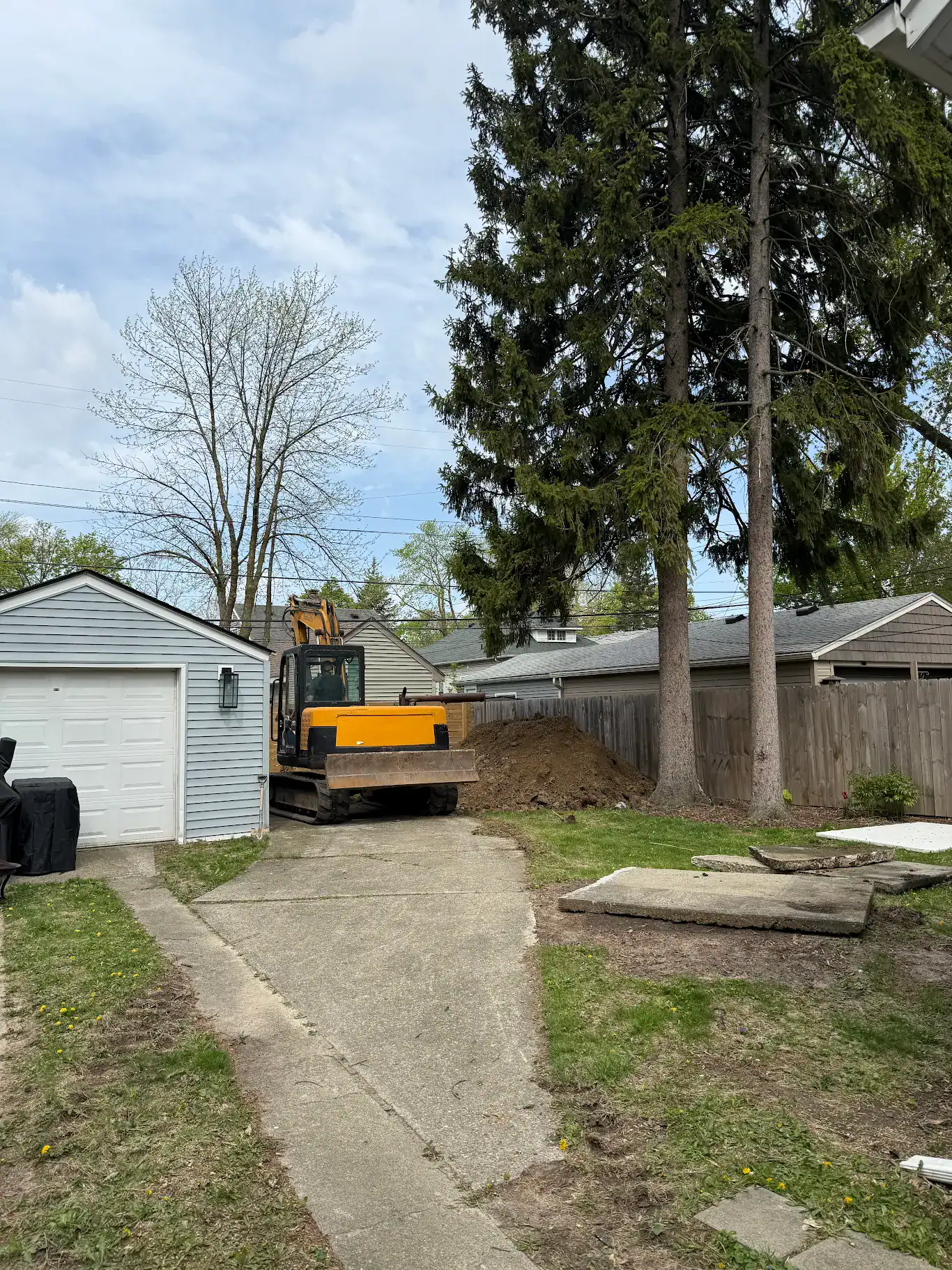 A mini excavator staged on a residential driveway at the start of an open-cut sewer line excavation in Metro Detroit