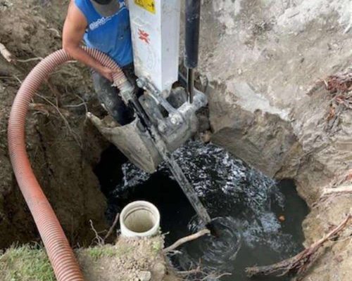 A person in a blue sleeveless shirt operates heavy machinery to pump dark, murky water from a deep hole—showcasing the expertise of bison plumbing warren michigan—with an orange hose and a white pipe visible nearby.