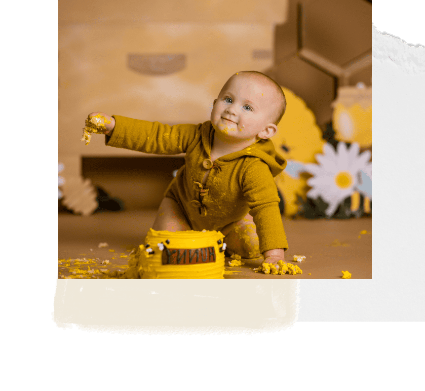 Colorful cake smash session with a smiling baby in mustard yellow outfit, celebrating a first birthday, with playful props and vibrant decorations.