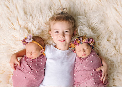 Adorable young girl with twin newborn sisters dressed in pink, lying on fluffy cream blanket, celebrating sibling bond and childhood innocence.