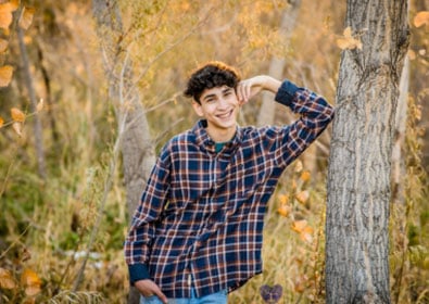 Urban fall portrait of a smiling teenage boy in a plaid shirt outdoors with trees and autumn leaves.