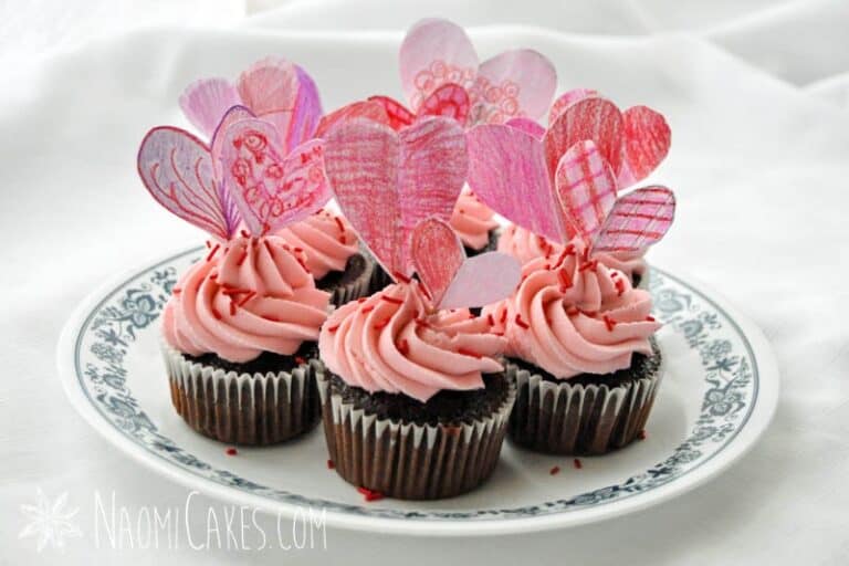 plate of chocolate cupcakes on white tablecloth with pink frosting, red sprinkles and crayon coloured hearts for Valentines Day