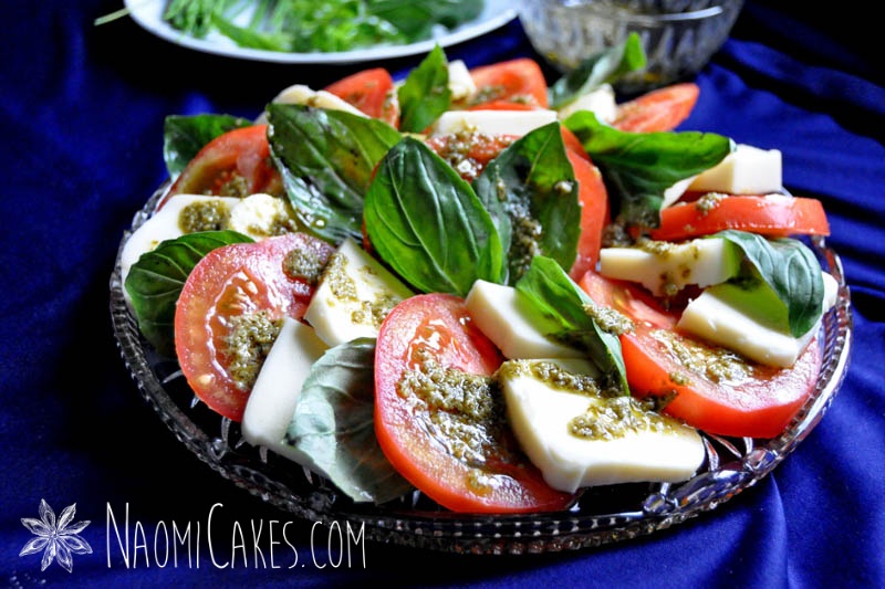tomatoes, pesto, fresh basil, and mozzerella on a round clear platter with a blue tablecloth and leftover ingredients in the background