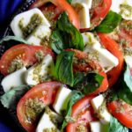 tomatoes, pesto, fresh basil, and mozzerella on a round clear platter with a blue tablecloth in the background