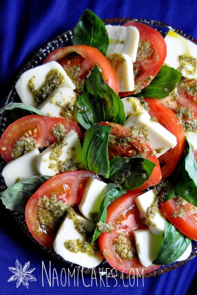 tomatoes, pesto, fresh basil, and mozzerella on a round clear platter with a blue tablecloth in the background