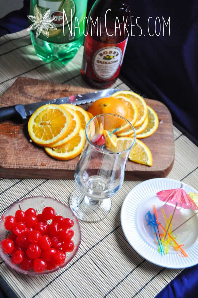 orange slices, maraschino cherries, empty glass, cocktail umbrellas, and sword toothpicks on a bamboo mat