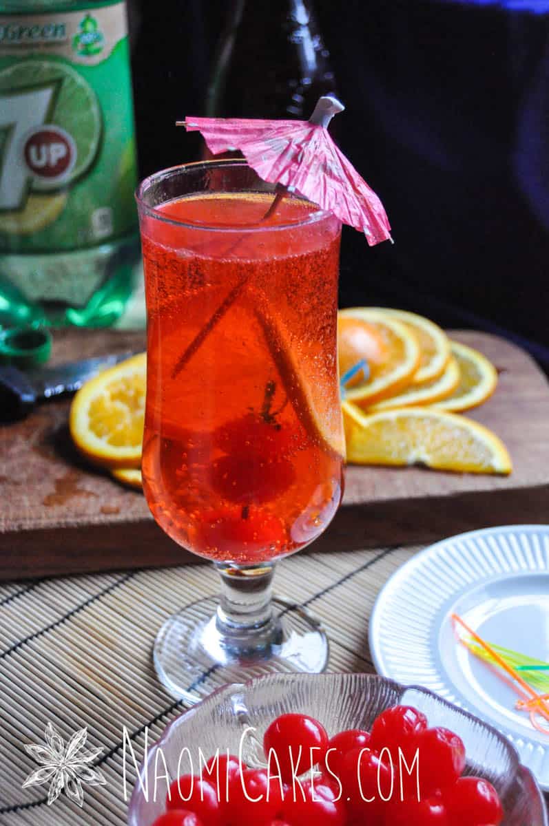 shirley temple mocktail in a long cup with a pink umbrella with orange slices in the background