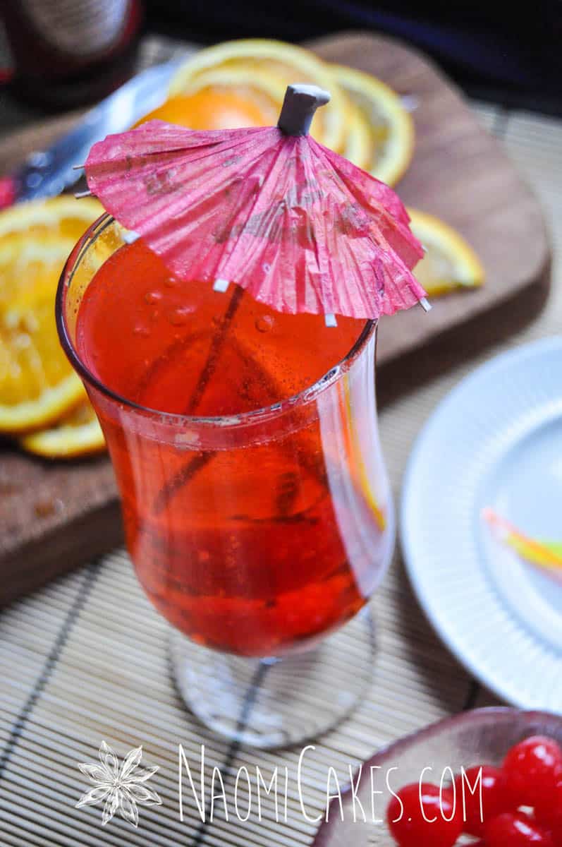 shirley temple mocktail in a long cup with a pink umbrella with garnishes in the background