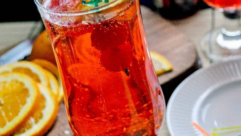 shirley temple mocktail in a long cup with a sword toothpick and yellow umbrella with garnishes in the background