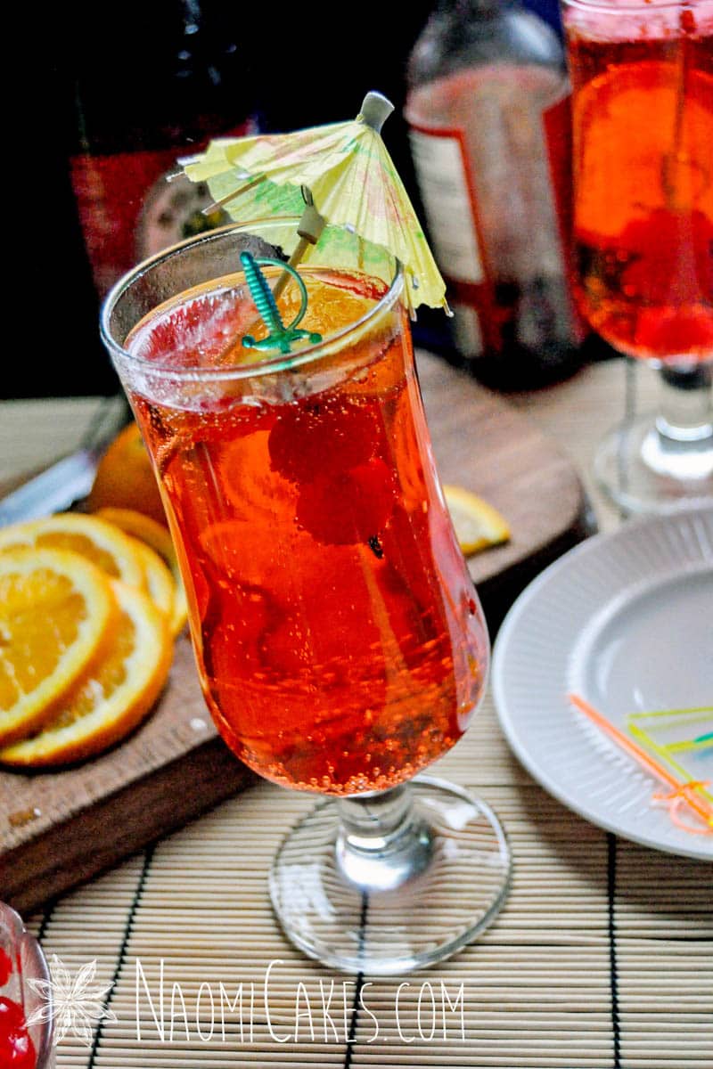 shirley temple mocktail in a long cup with a sword toothpick and yellow umbrella with garnishes in the background