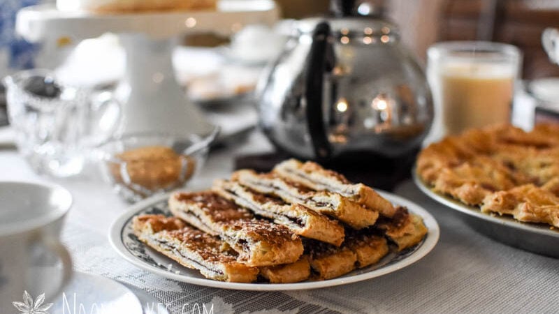 plate of cinnamon sprinkled baked pastry on a table with a lace tablecloth, a silver tea pot and cake pedestal in the background