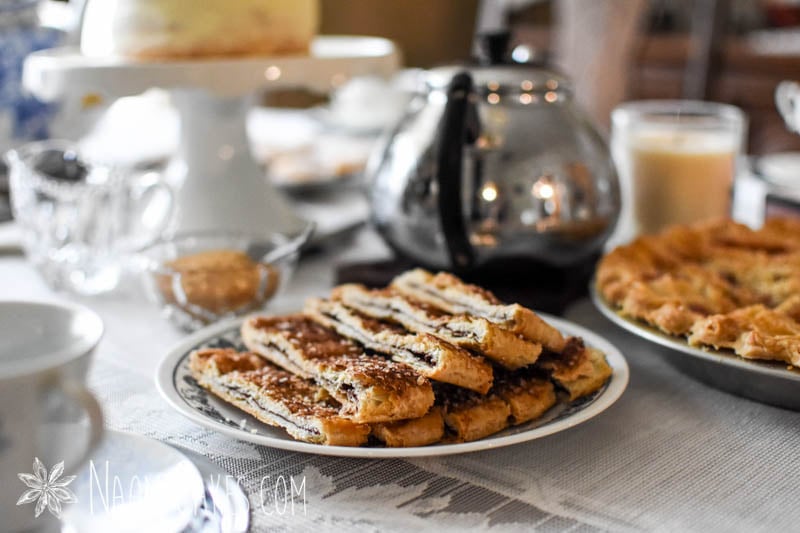 plate of cinnamon sprinkled baked pastry on a table with a lace tablecloth, a silver tea pot and cake pedestal in the background