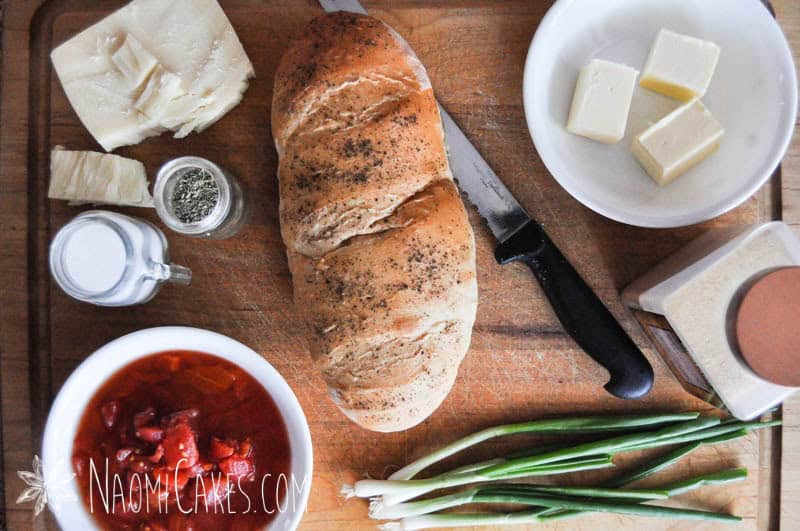 bread, cheese, green onions, tomatoes, and seasonings on a cutting board with a knife