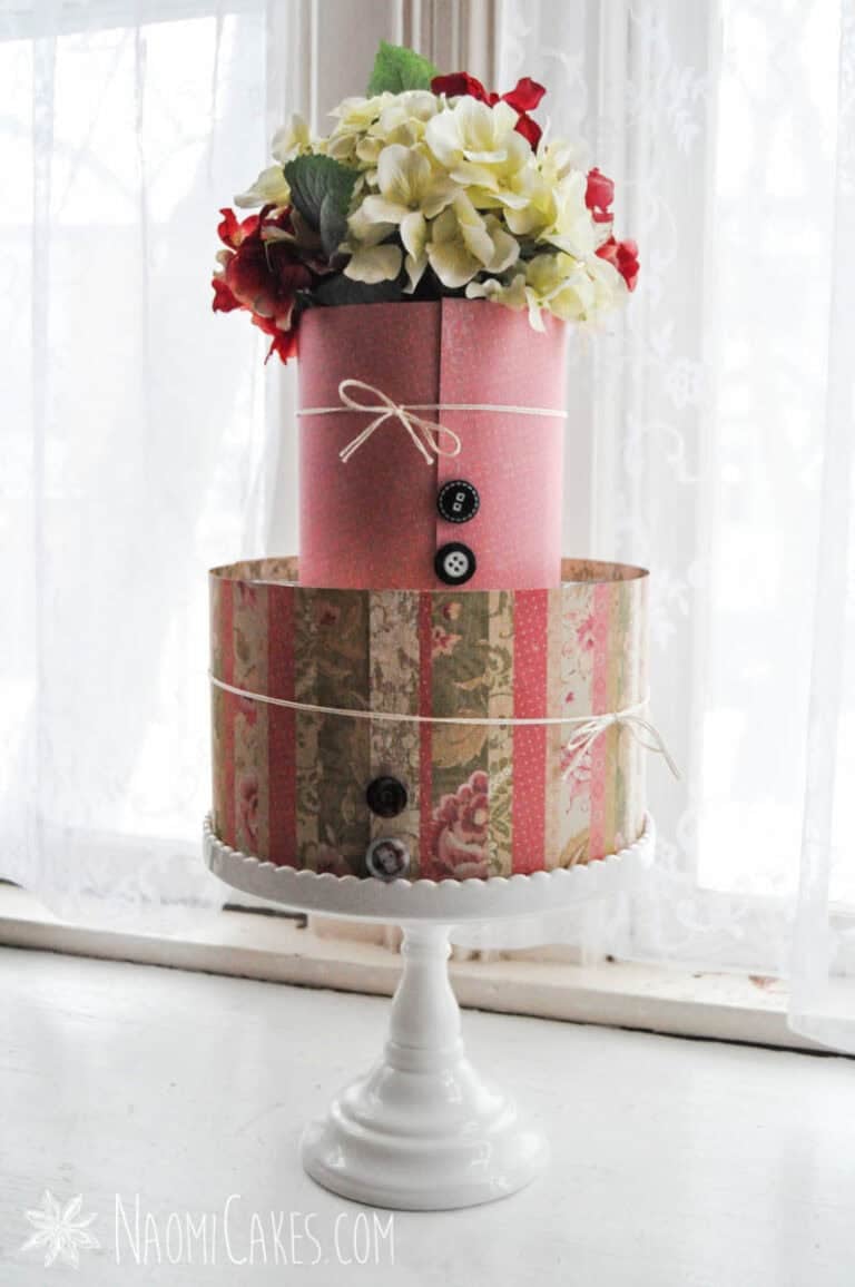 2 tiers of foam cake dummies wrapped in pink and floral scrapbooking paper, topped with silk hydrangias, fastened with button thumb tacks, and tied with string