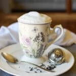 a foamy tea latte in a tall china tea cup with lavender and roses on a plate with a tea ball and spoon and a white cloth