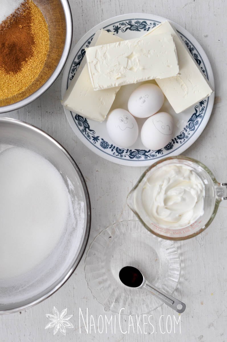 sugar, eggs with faces on them, vanilla, graham cracker crumbs, cream cheese and sour cream on a white table