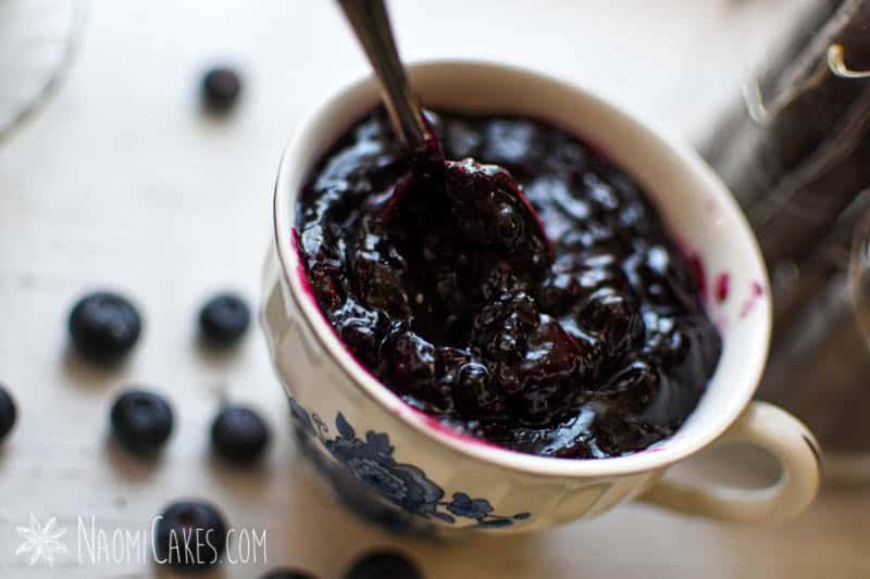 a teacup full of blueberry lemon pie filling with a spoon in it and scattered fresh blueberries around on the white table