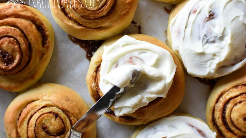 Some unfrosted and some frosted cinnamon rolls on a pan, with a knife spreading icing on one