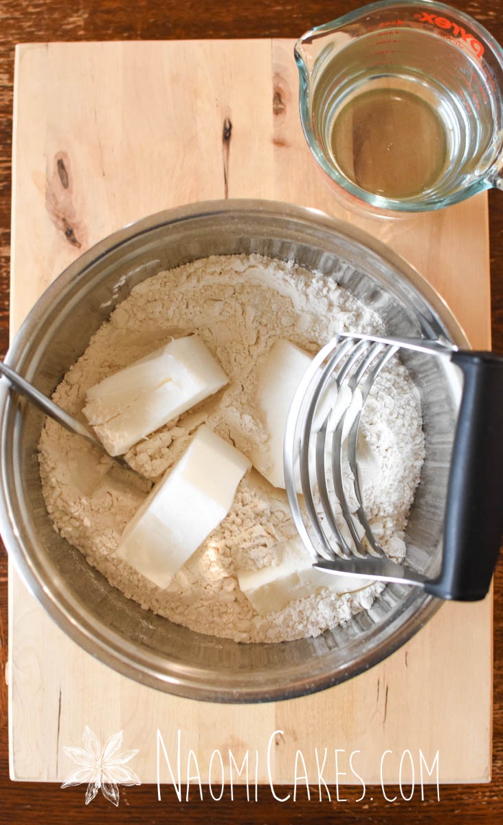 bowl with flour, shortening, and a pastry cutter on a board with a measuring pitcher of water