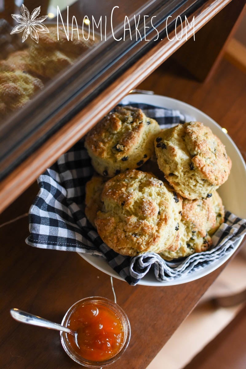 bowl of currant scones reflecting in a glass display case with a checkered tea towel and orange jam