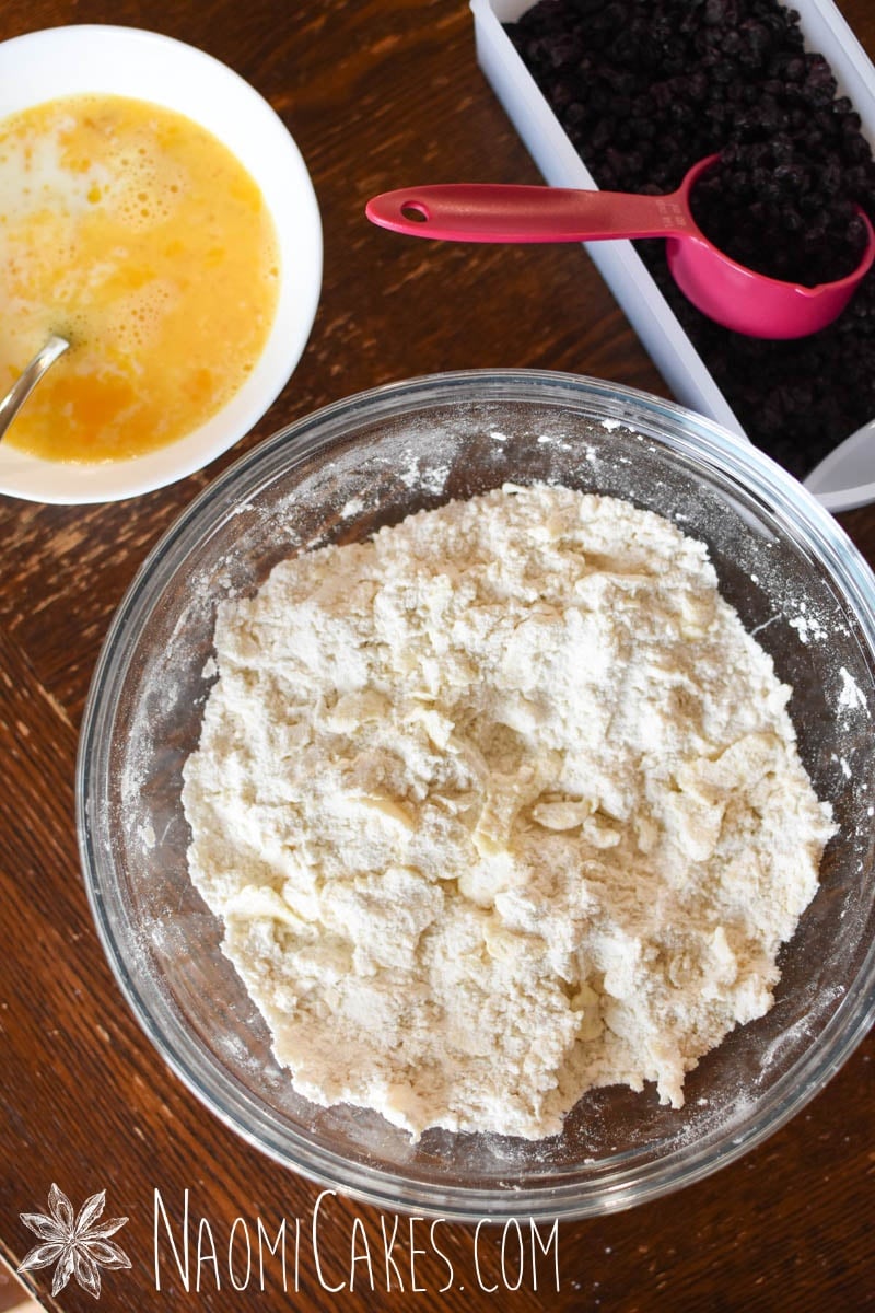flour and butter mixed together with a bowl of egg and milk and currants on the table beside it