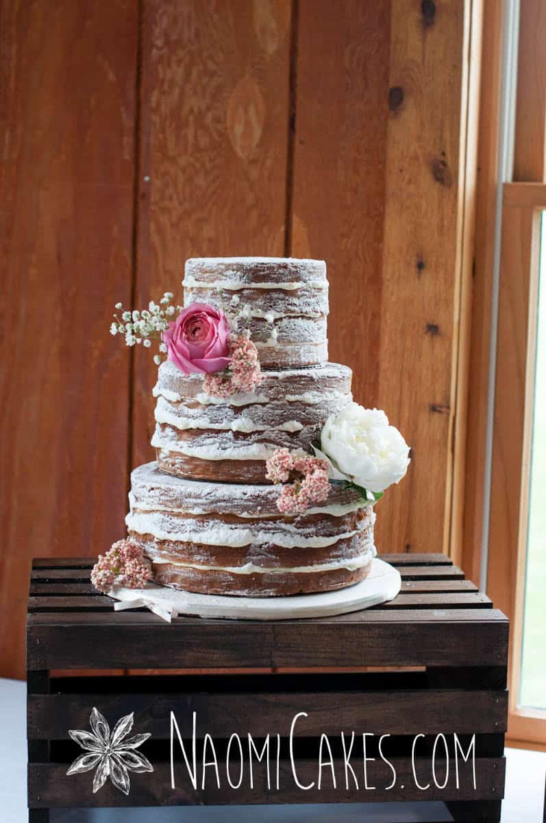 full image of a naked cake wedding cake with pink flowers on a wooden crate with log cabin background