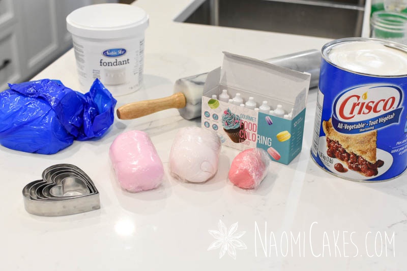fondant in shades of pink, a rolling pin, a bucket, Crisco shortening, and food colouring on a white countertop