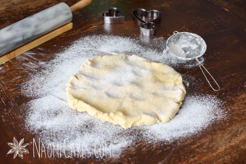 sugar cookie dough on a wooden table with heart cutters, a sieve, and a rolling pin