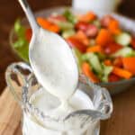 spoon dripping into a glass pitcher of creamy yogurt salad dressing with a garden salad