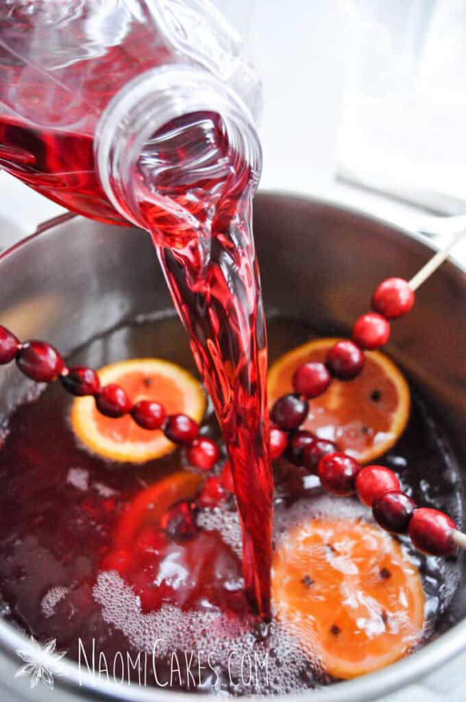 cranberry juice being poured over cranberries and citrus with spices into a pot with apple cider
