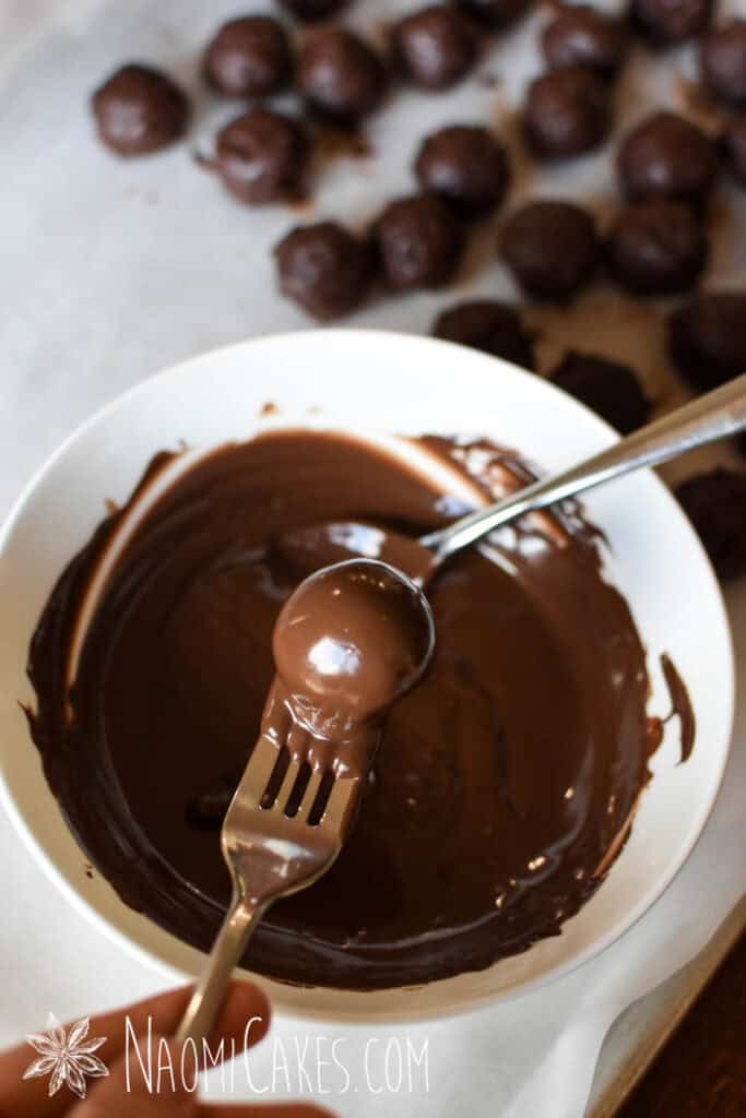 a chocolate coated dark chocolate truffle on a fork above a white bowl full of melted chocolate and a spoon