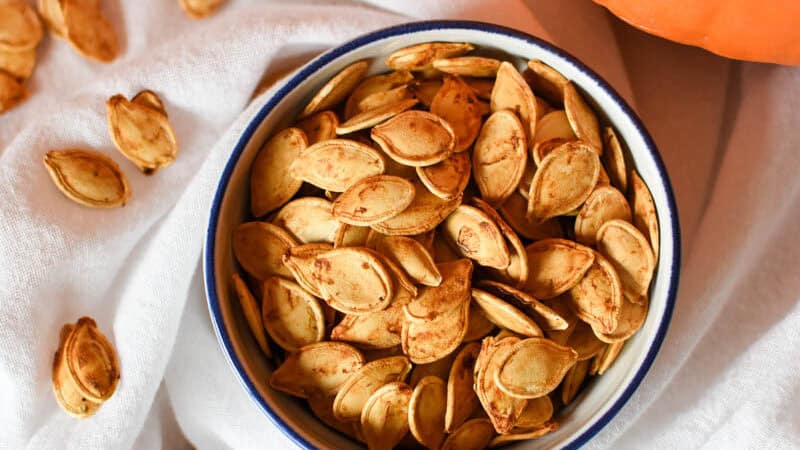 overhead view of a bowl of roasted pumpkin seeds with seeds scattered around and pumpkins in the background