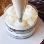 a piping bag and star tip piping swirls of whipped cream onto a layered chocolate cake with a plastic collar around it