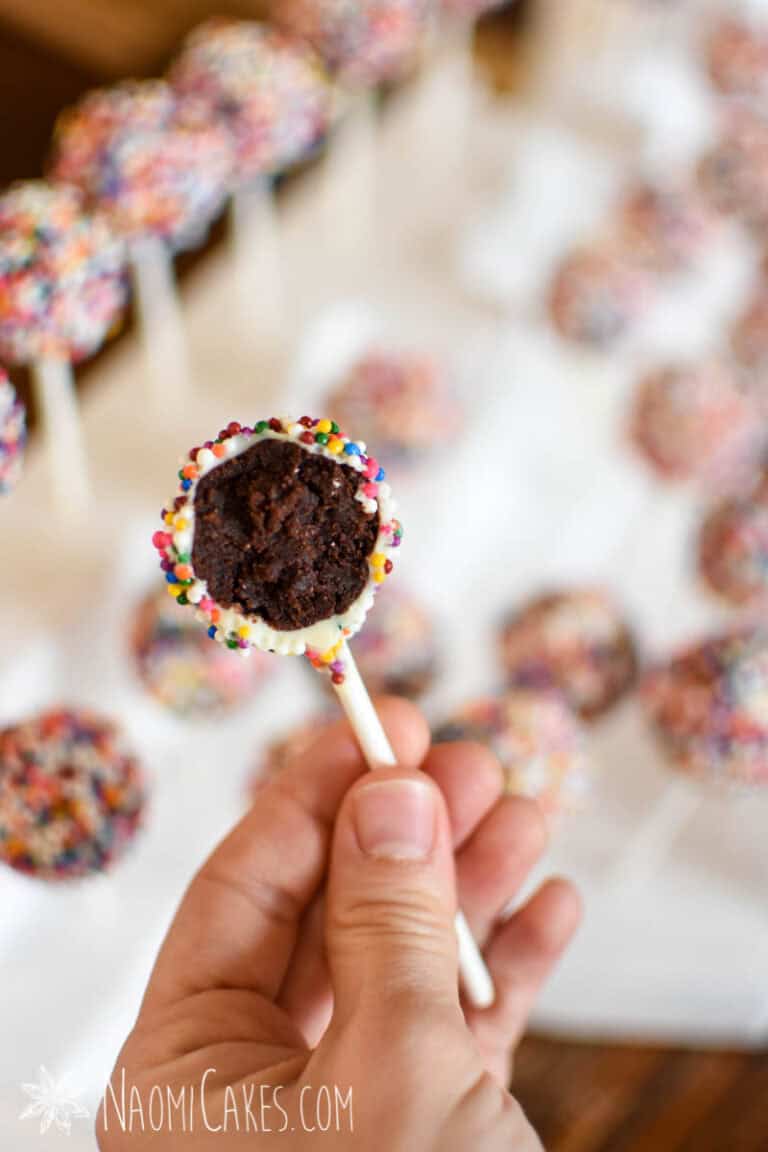 a hand holding a sprinkle covered chocolate cake pop with a bite taken out to show in the inside