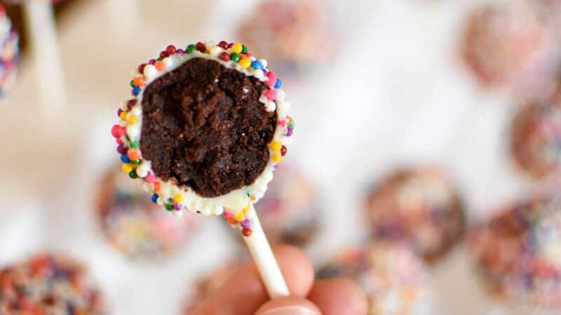 a hand holding a sprinkle covered chocolate cake pop with a bite taken out to show in the inside