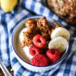 close up of a ramekin with yogurt topped with granola, raspberries and banana slices with more granola and bananas in the background on a blue gingham cloth