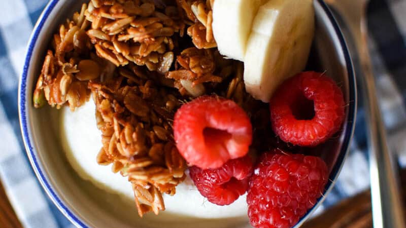 top view of a ramekin with yogurt topped with granola, raspberries and banana slices on a blue gingham cloth