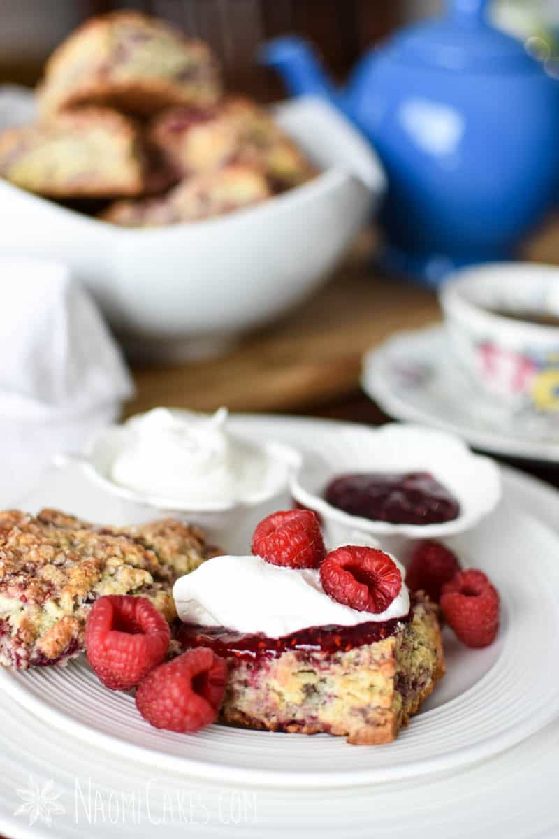 raspberry scone on a white plate with raspberry jam, cream, fresh berries, and a blue tea pot, a tea cup, and more scones in the background