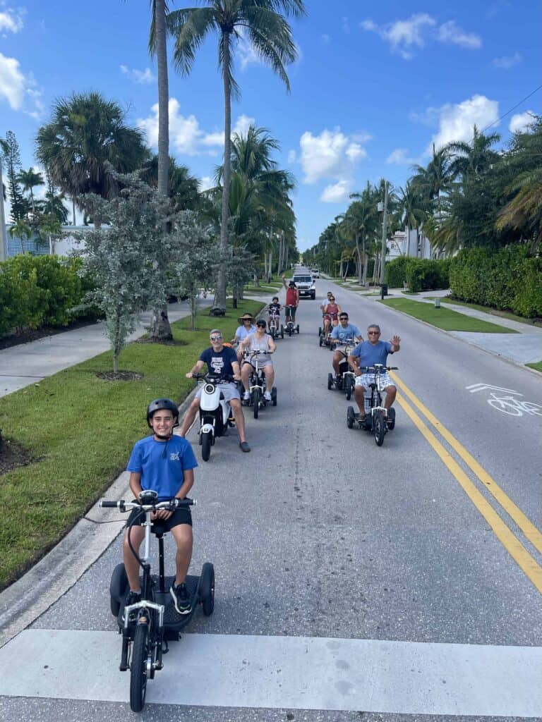 A group riding electric trikes and mopeds together along a palm-lined street in Naples