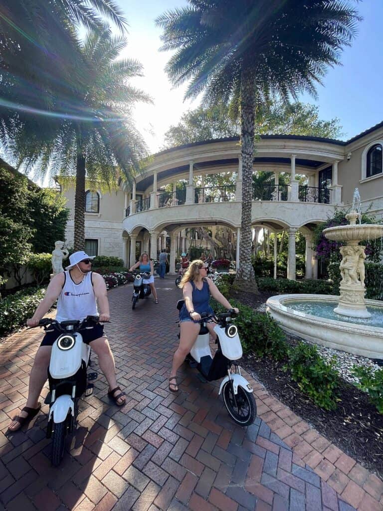 Electric moped riders beside a fountain and a grand Naples coastal estate