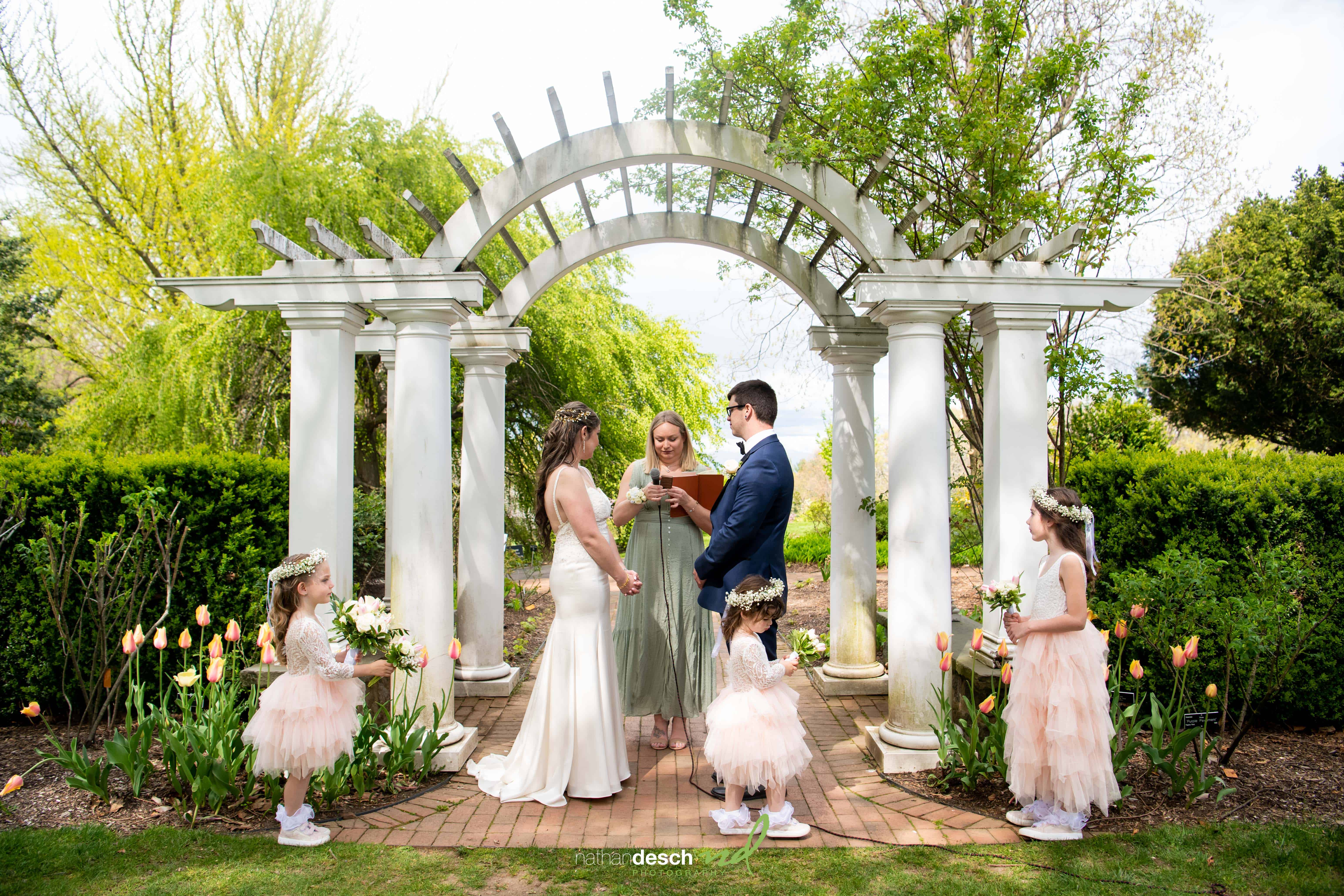 Bride and groom, Billy and Kaitlyn, exchange vows under a white garden arch with flower girls in spring wedding ceremony.