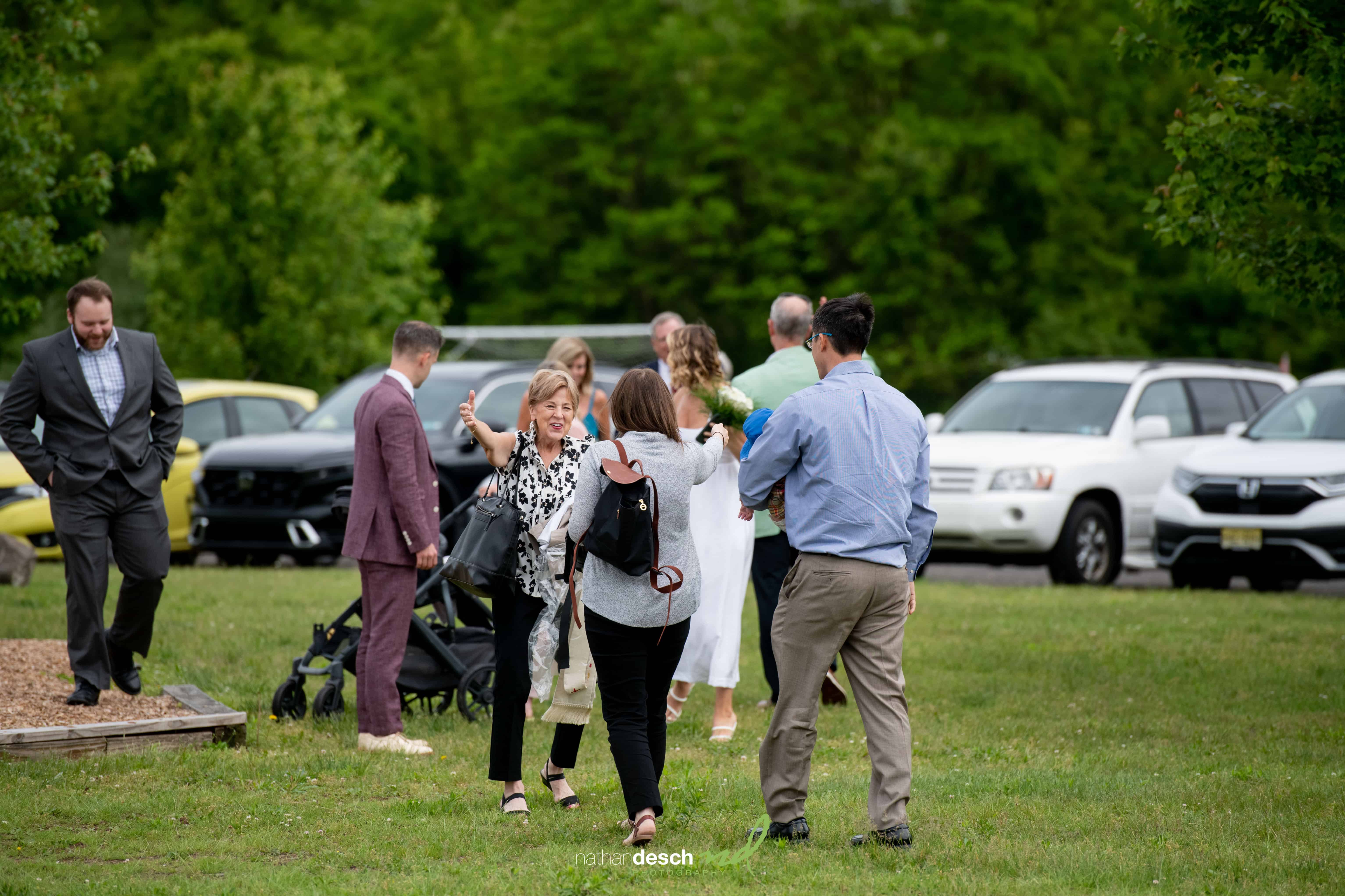 Wedding guests greeting each other on grassy lawn with trees, parked cars, and a smiling woman waving in the foreground.