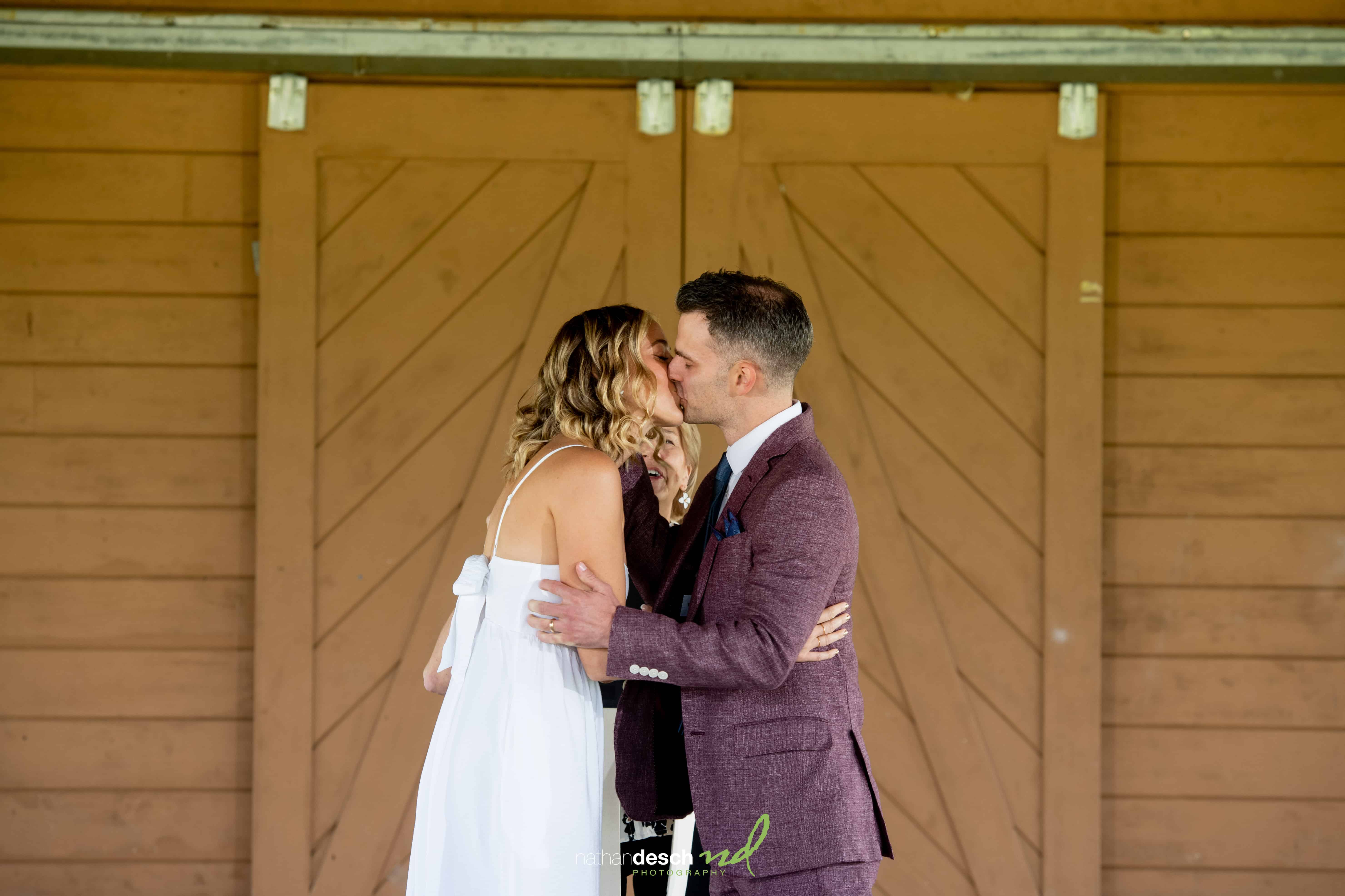Bride in white dress kisses groom in maroon suit jacket before large brown wooden doors on wedding day.