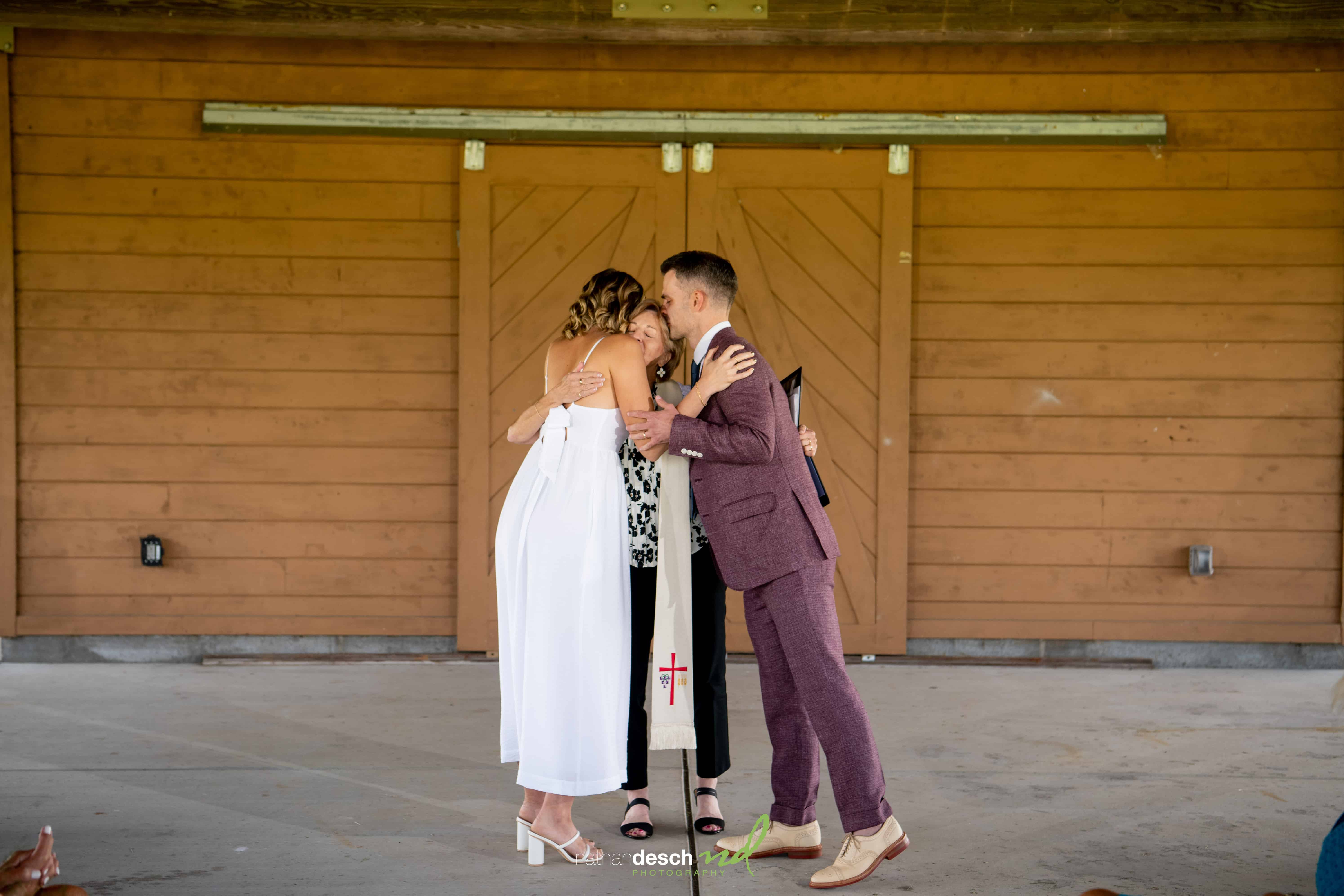 Bride and groom hugging officiant in front of wooden doors during wedding ceremony, bride in white dress, groom in purple suit.