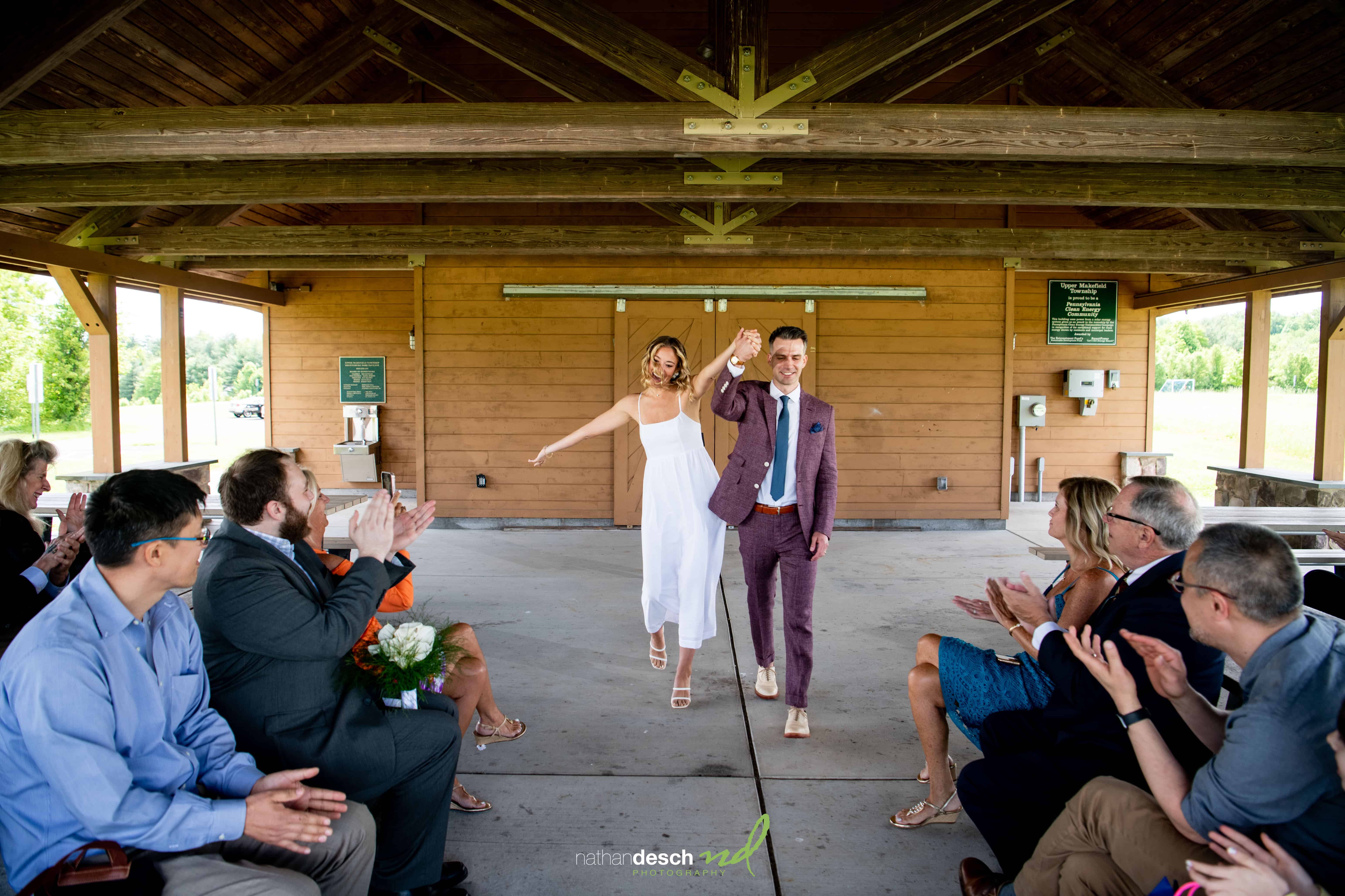 Bride and groom walk hand in hand down aisle under pavilion as wedding guests cheer, sunlight streaming in.