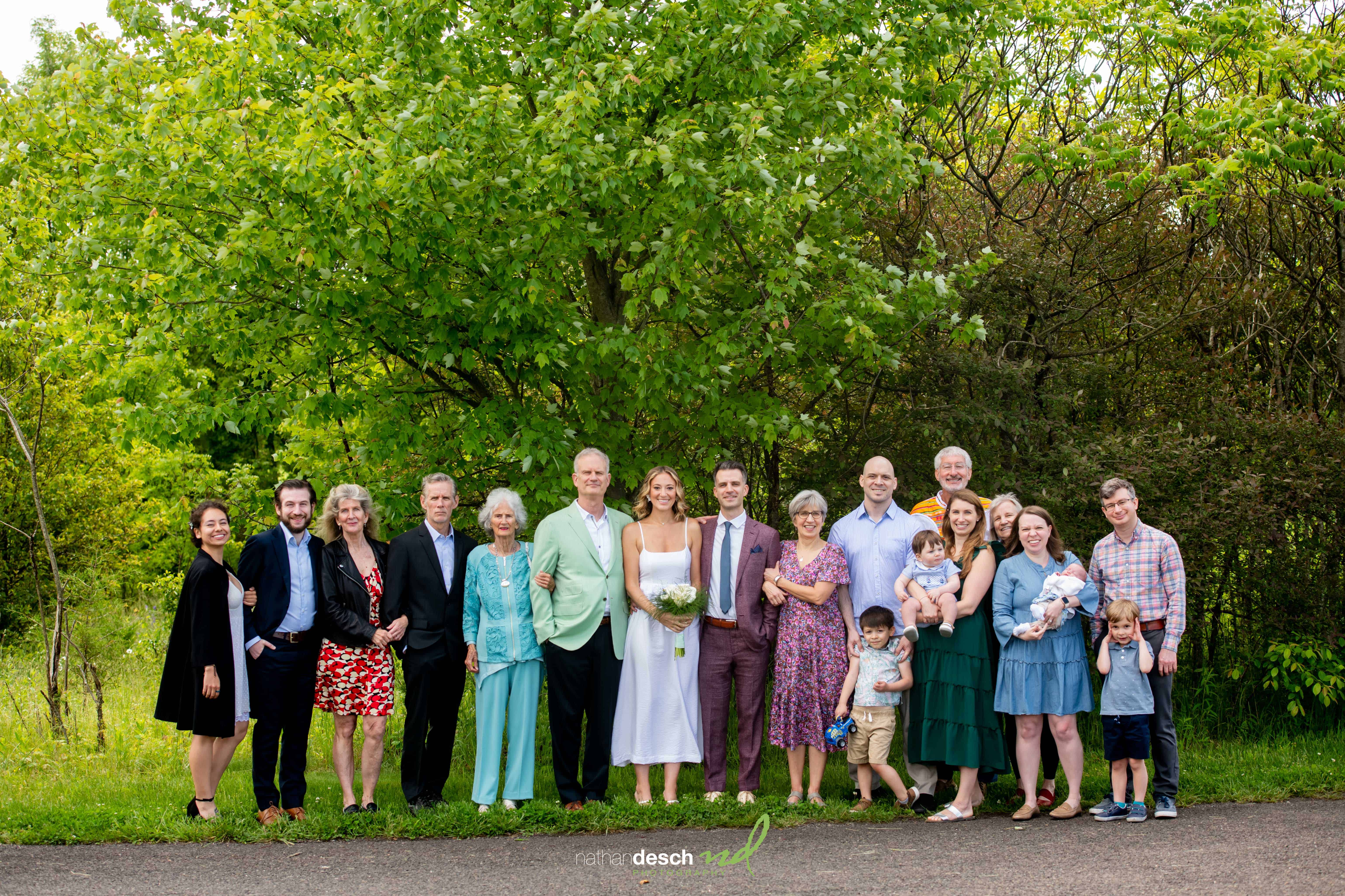 Wedding party smiling outdoors in front of green trees, with adults, seniors, and children in semi-formal attire.