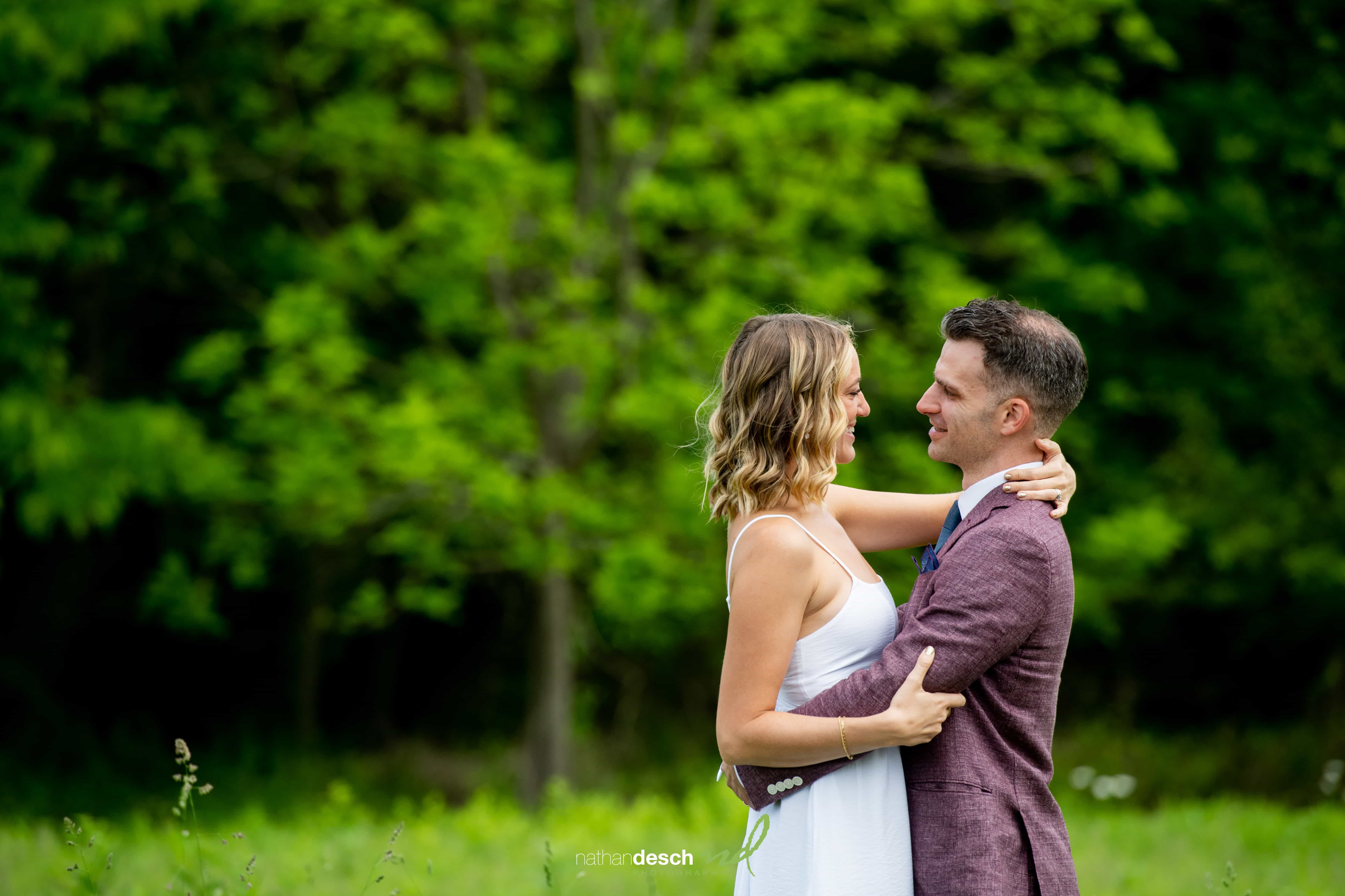 Bride and groom smiling and embracing outdoors with green trees, bride in white dress, groom in rust suit and blue tie.