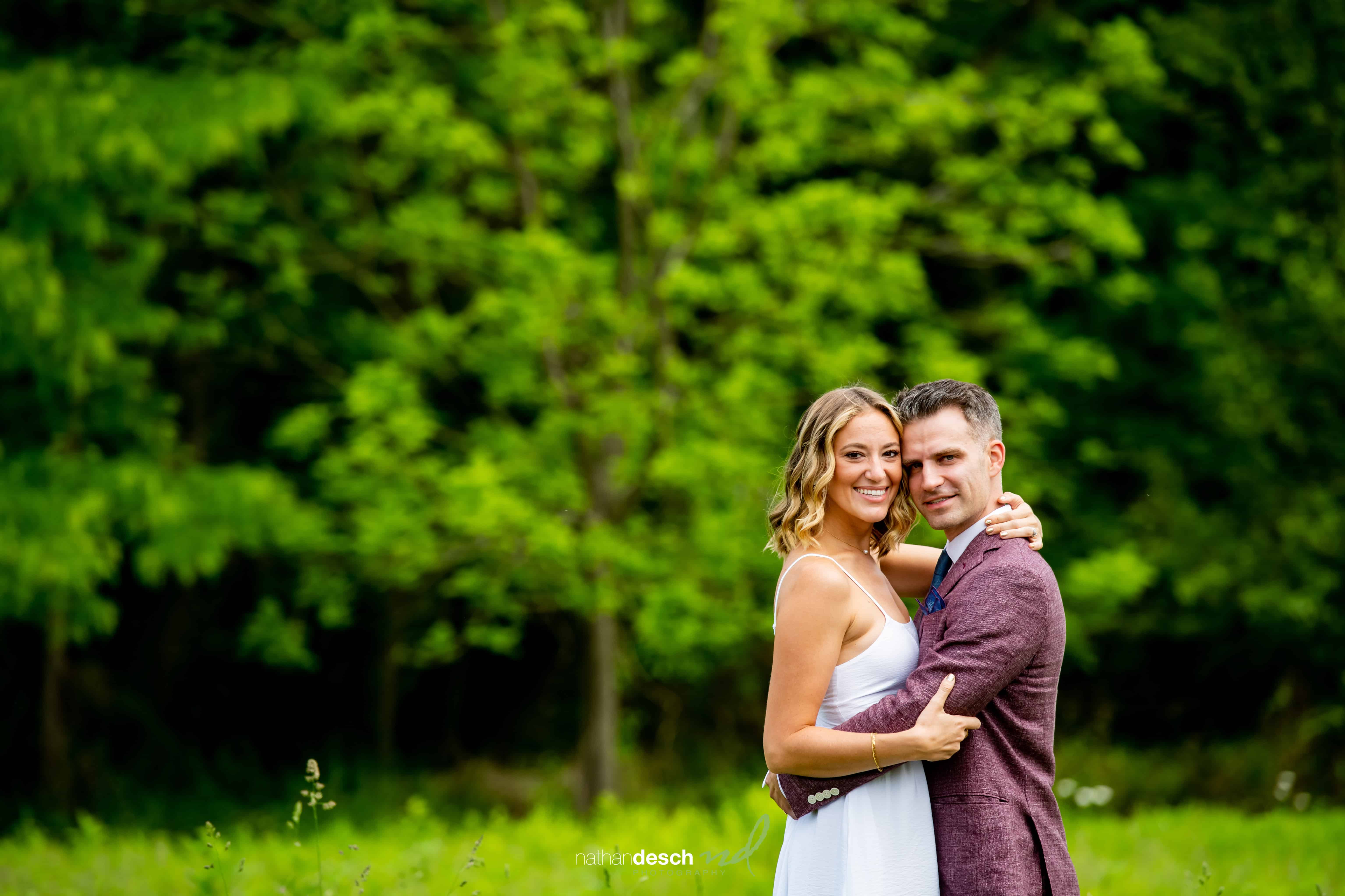 Bride and groom smiling and embracing outdoors with lush green trees and grass on a sunny wedding day.