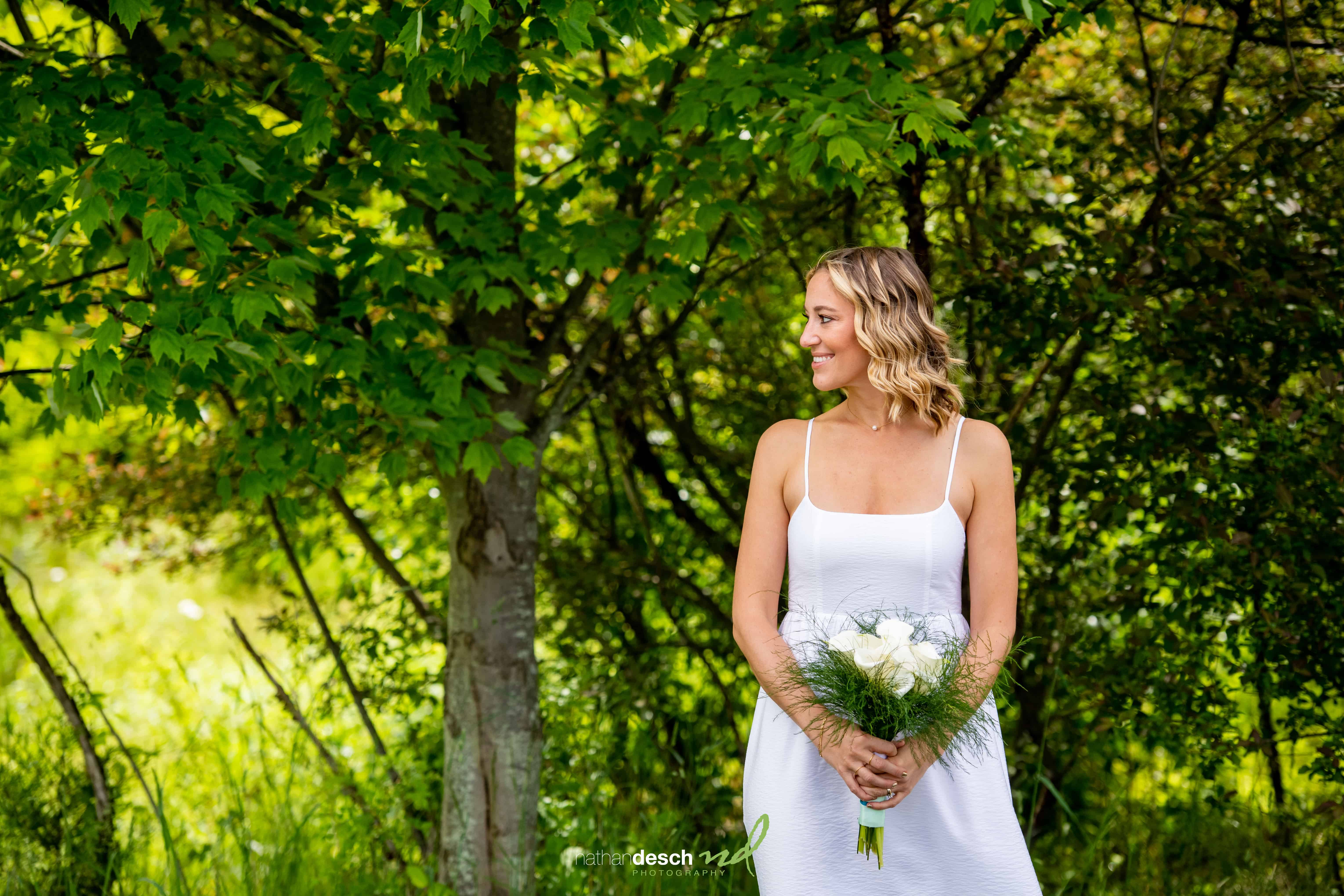 Bride in white dress holding bouquet, smiling under sunlit trees at Will and Laura’s outdoor wedding ceremony.